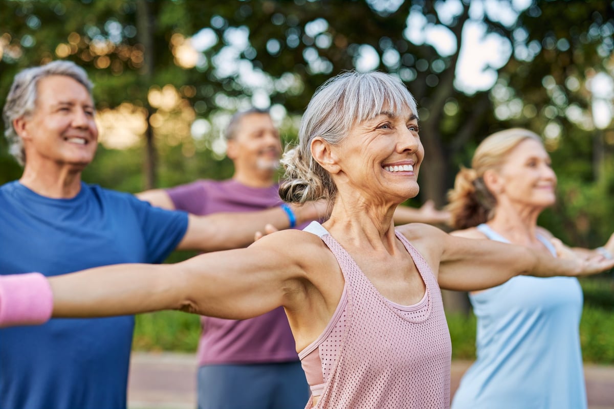 Happy senior group doing yoga together in the park, gentle gymnastics. Mature men and women stretching arms in outdoor fitness class.