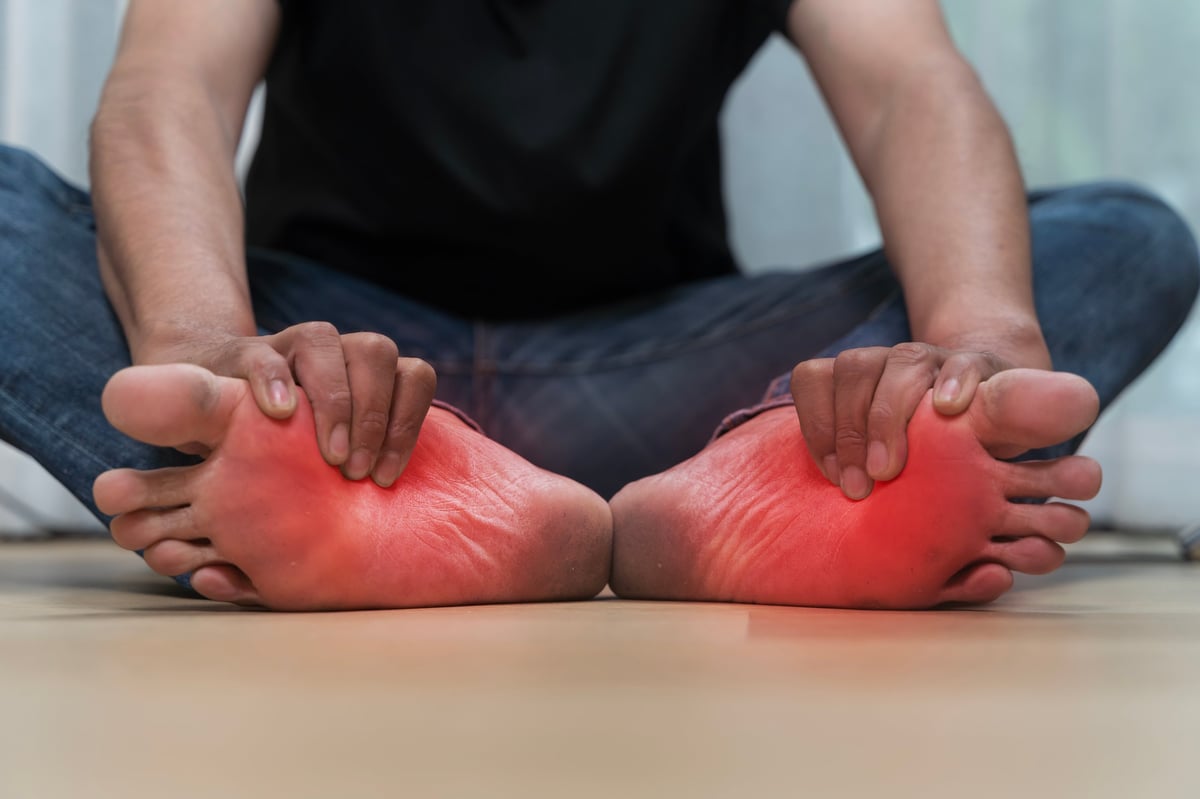Close up of man sitting on floor holding both feet with visible redness and pain, symptoms of inflammation, plantar fasciitis, neuropathy, gout, or diabetic foot problems related to chronic conditions