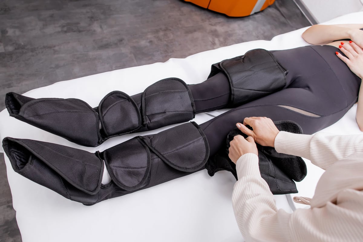 Therapist Operating Pressotherapy Machine While Female Client Lies with Full Leg Air Compression Boots on Massage Table — Professional Pneumatic Lymphatic Drainage Treatment for Legs, Circulation Therapy and Modern Aesthetic Medicine Spa Concept