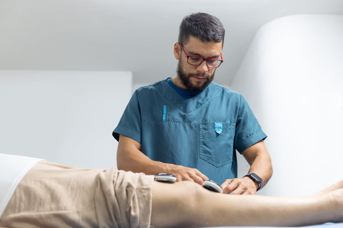 Professional physiotherapist performing electrotherapy treatment on a patient's knee using electrical stimulation devices during a rehabilitation session in a modern medical clinic.