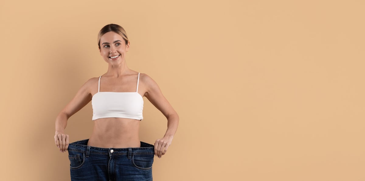 Young beautiful woman smiling while holding loose jeans after weight loss, representing fitness, body confidence and healthy lifestyle