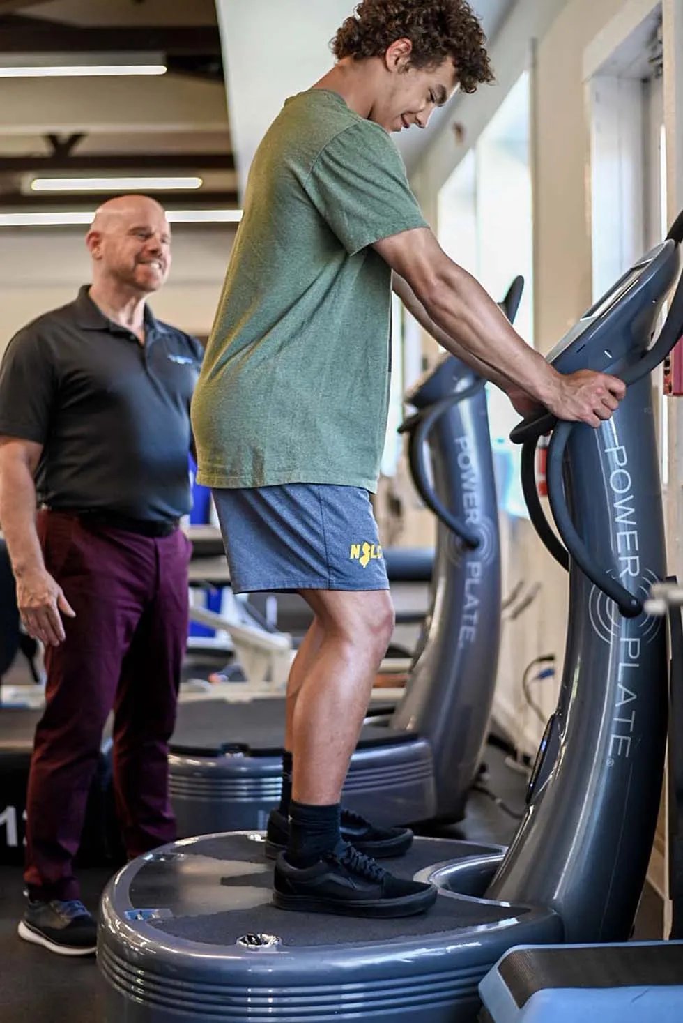 Man using a vibration plate machine in a gym while a trainer in dark shirt watches and supervises